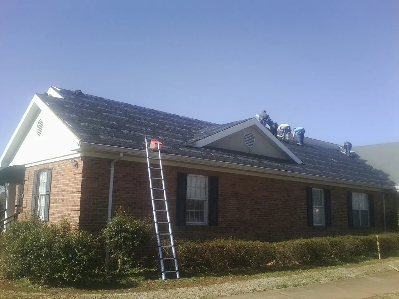 Crew tearing shingles off a brick ranch roof with a ladder set — Emergency Roof Repair work by our crew in Whitmore Lake