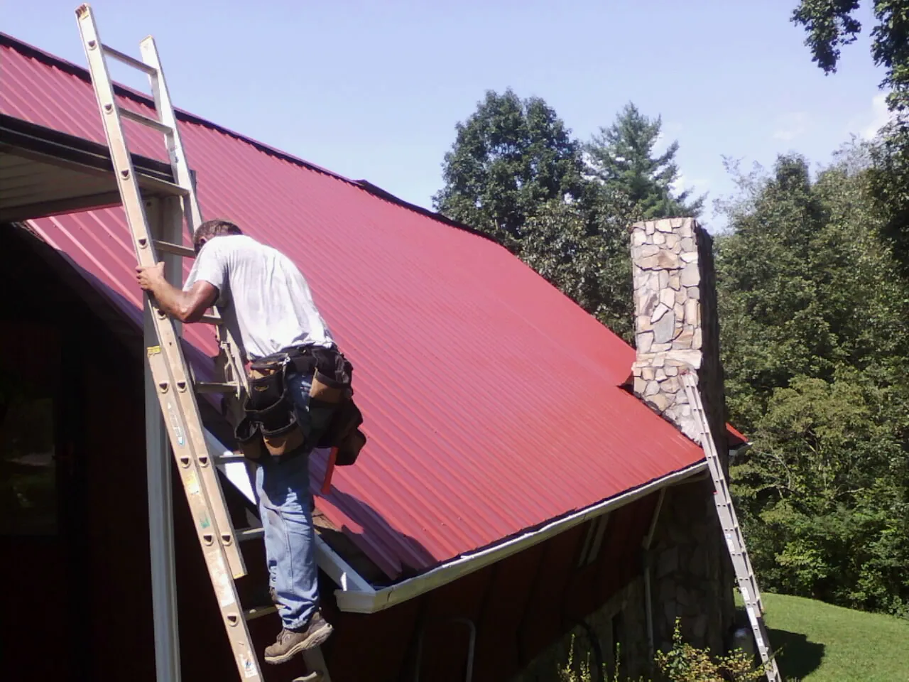 Roofer on a ladder beside a red metal roof and stone chimney — Soffit & Fascia Repair by Roofing Brothers in Whitmore Lake